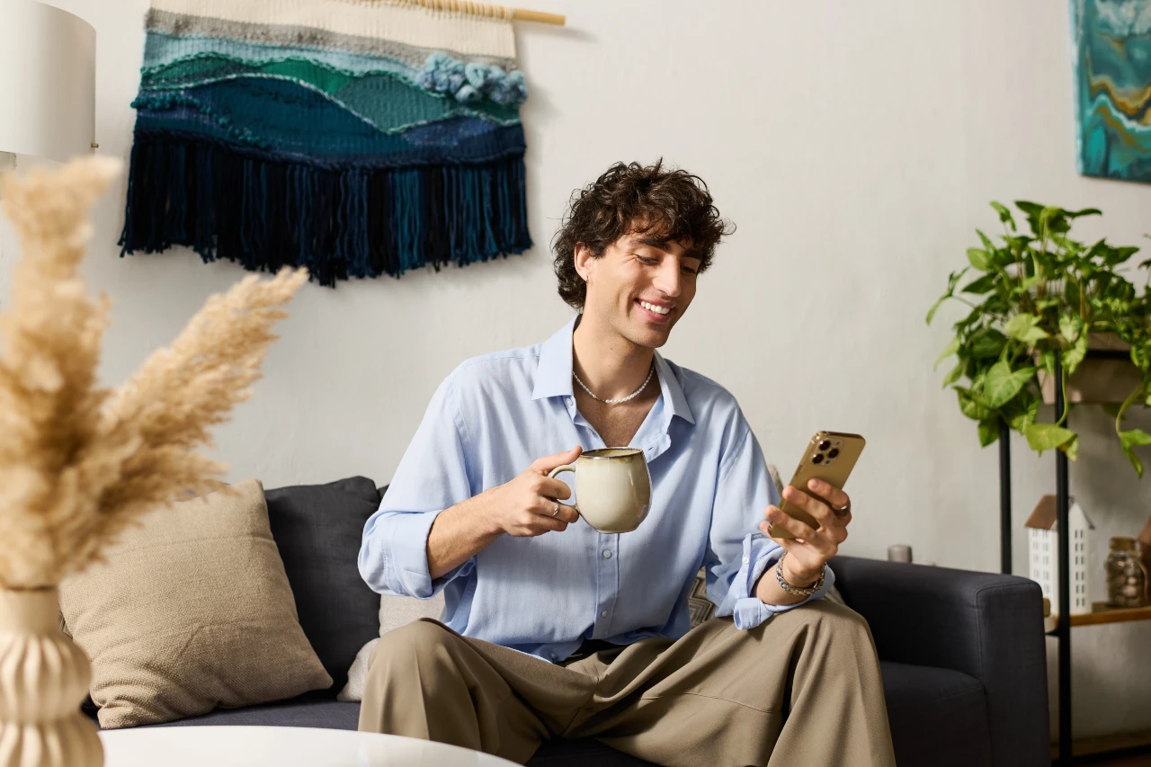 Der Dresscode Casual Chic ist lässig aber schick zugleich. Dresscode Cacual Chic, Mann sitzt mit einer Tasse Kaffee auf der Couch und tägt ein hellblaues, lockeres Hemd und eine Stoffhose