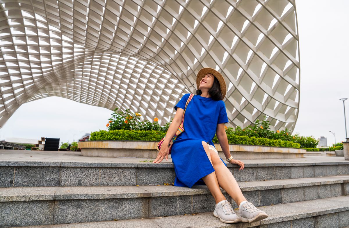 Geschlossenes blaues Kleid, sommerlich dank Schlitz und kurzen Ärmeln trägt eine Frau auf einer Treppe mit Sommerhut