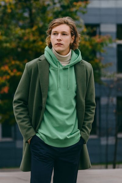 Junger Mann trägt mintgrünen Oversized-Hoodie unter olivgrünem Mantel, herbstlicher Layering-Look im urbanen Umfeld.