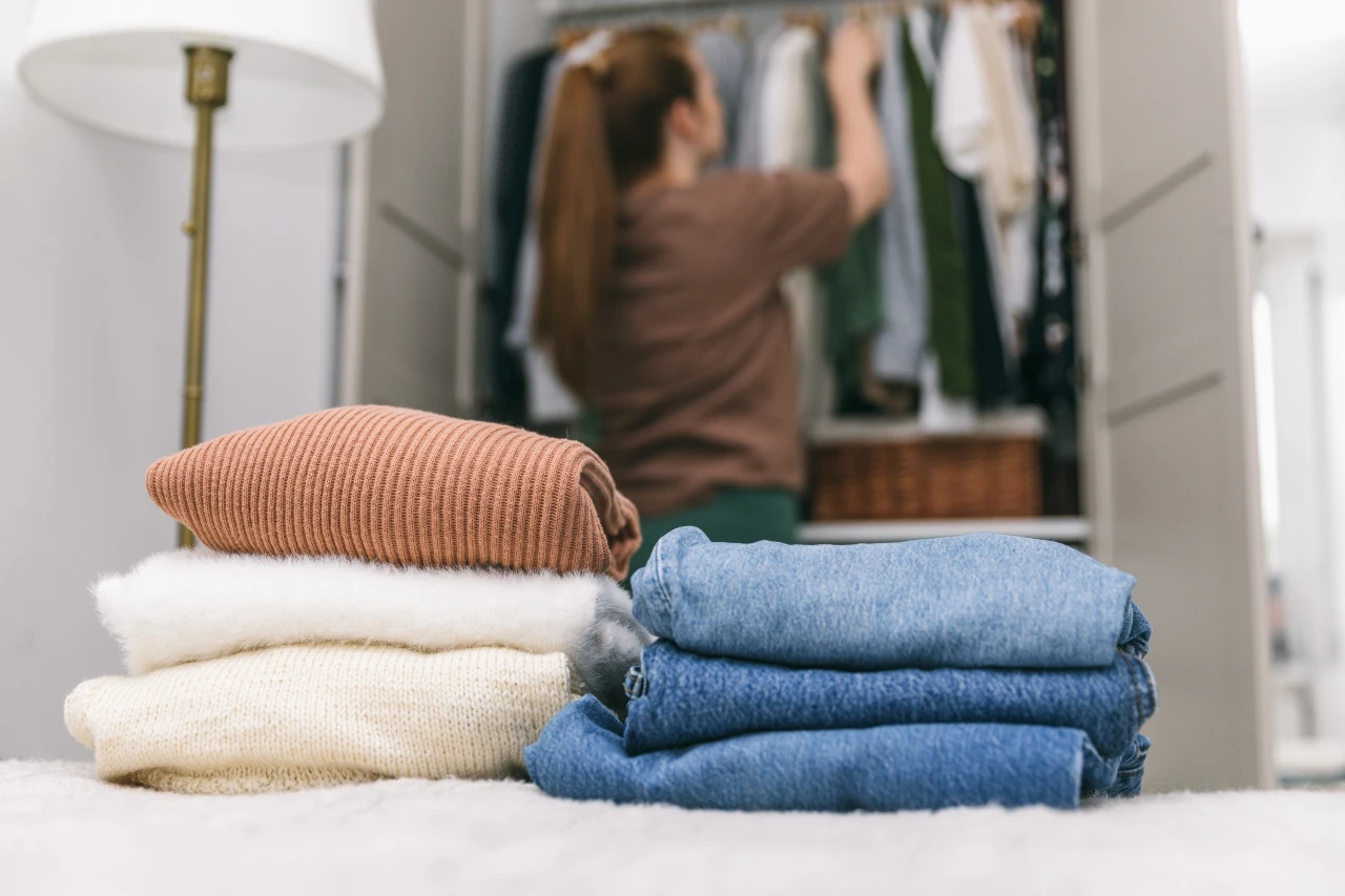 Schritt für Schritt zum minimalistischen Kleidungsstil Frau räumt Kleiderschrank aus, hinter ihr liegen Stapel mit Jeans und Pullover