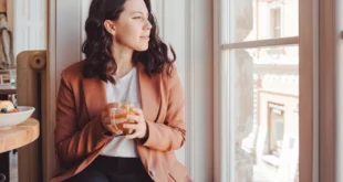 Frau in schwarzer Jeans, weißem Shirt und rostbraunem Blazer sitzt am Fenster und schaut heraus