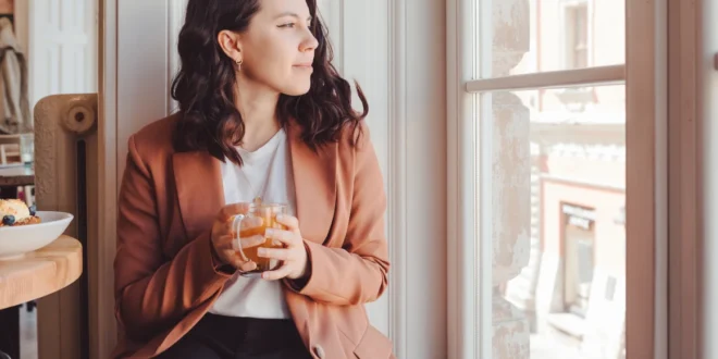 Frau in schwarzer Jeans, weißem Shirt und rostbraunem Blazer sitzt am Fenster und schaut heraus