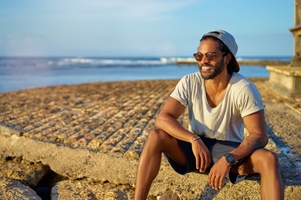 Ein junger Mann sitzt am Strand. Er trägt eine Sommercap mit dem Schirm nach hinten. Coole Sommercaps