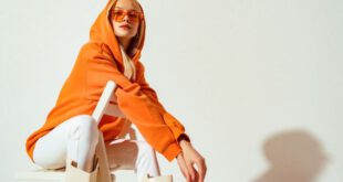 Stylische Frau mit orangenem Hoodie, Sonnenbrille, weißer Jeans und langen Stiefeln in weiß