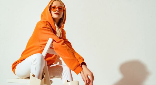 Stylische Frau mit orangenem Hoodie, Sonnenbrille, weißer Jeans und langen Stiefeln in weiß