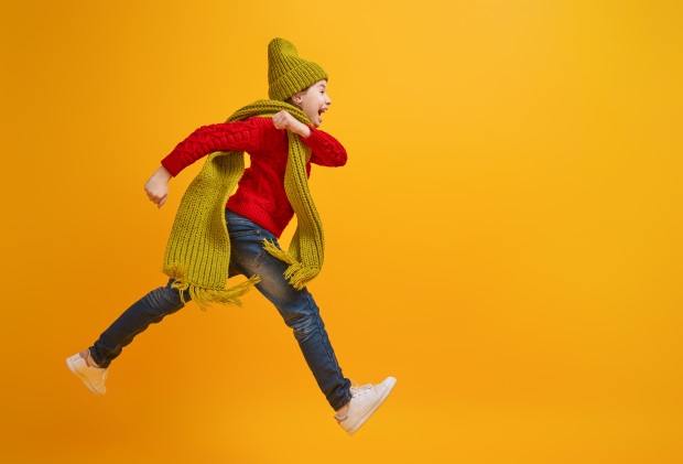Ein kleiner Junge, gekleidet mit einem roten Wollpulli, blauer Jeans, gelbem Schal und gelber Mütze, springt ein Stück in die Luft. Kontrastfarben liegen im Trend und lassen sich vielfältig kombinieren. 