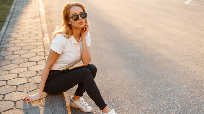 Frau in weißem Poloshirt und schwarzen Jeans sitzt am Straßenrand und schaut in die Kamera