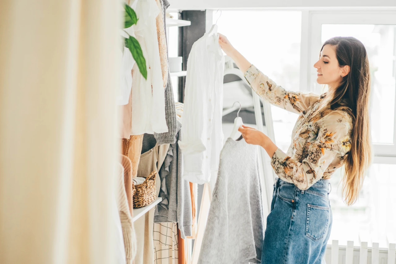 Frau steht vor dem Kleiderschrank und hat zwei Kleidungsstücke in der Hand und lächelt.