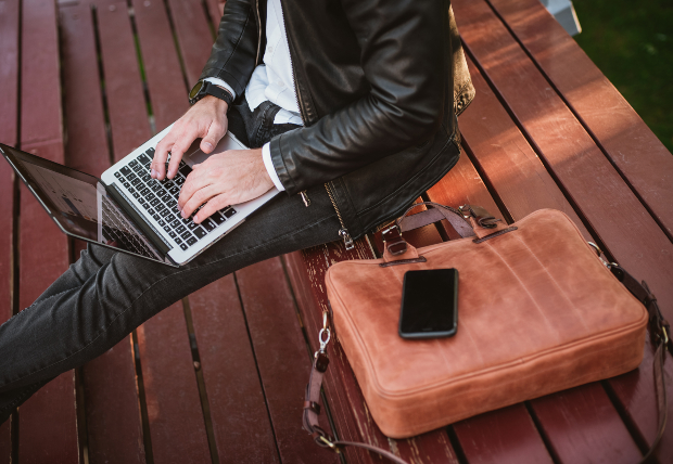 Ein Mann sitzt auf einer Bank auf benutzt sein Laptop, neben Ihm liegt eine schicke Notebooktasche