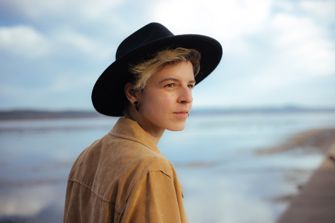 Androgyne Person mit Hut, Ohrring und beiger Lederjacke