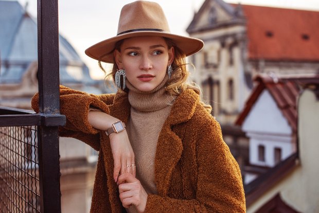 Eine elegante Frau in einem beigen Mantel posiert vor einer historischen Stadtkulisse