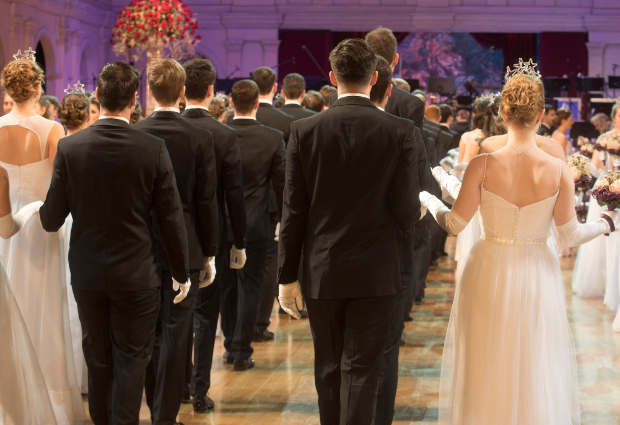 Auf einer Ballveranstaltung schreiten mehrere Männer im Frack und Damen im Abendkleid in einen Saal.