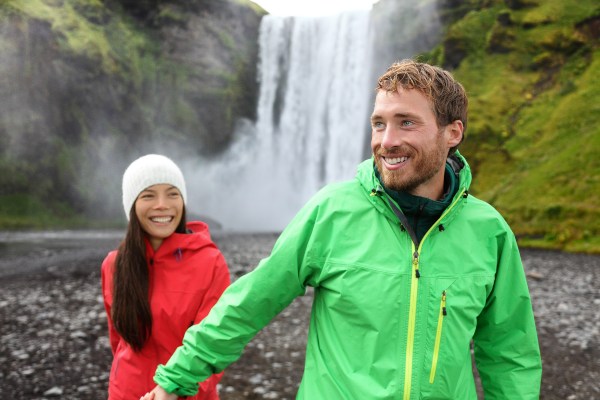 Ein junges Paar bei unbeständiger Witterung vor einem Wasserfall, sie tragen Hardshelljacken Jacken-Material