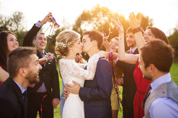 Brautpaar küsst sich, Gäste stehen darum herum - Festliches Outfit ist bei Hochzeiten Pflicht