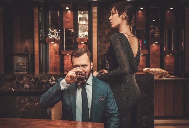 Junges Paar, Mann sitzt und trinkt Alkohol, Frau in elegantem Kleid steht - Festliches Outfit