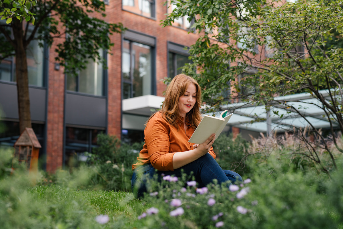 Eine Bluse ist ein unverzichtbares Basic beim Business Outfit. Frau mit Business Casual Outfit mit orangener Bluse und blauen Jeans sitzt auf Wiese und liest