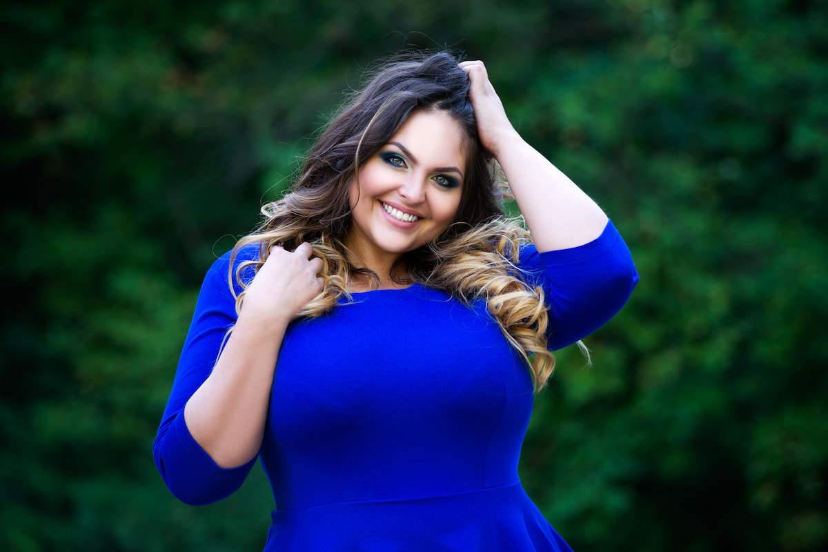 Stilvolle Schnitte und liebevolle Details betonen die natürliche Figur – ob mit Dreiviertelärmeln, feinen Säumen oder fließenden Stoffen Curvy Frau mit lockigen Haaren und blauem Dreiviertel-Ärmel-Oberteil lächelt in die Kamera